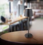 Die Tischleuchte - Vannie, eine elegante und moderne Tischlampe von Licht Factory mit zylindrischem Sockel und konischem Schirm, steht elegant auf einem Holztisch. Der Hintergrund zeigt eine Café-Kulisse mit hohen Fenstern und Thekensitzen in weichem Fokus.