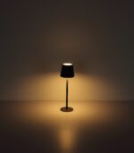 Die Tischleuchte - Vannie von Licht Factory ist eine moderne Stehleuchte mit dunklem Schirm, die ein warmes, sanftes Licht abgibt. Zentral vor einem schlichten Hintergrund platziert, schafft sie eine ruhige und minimalistische Atmosphäre.