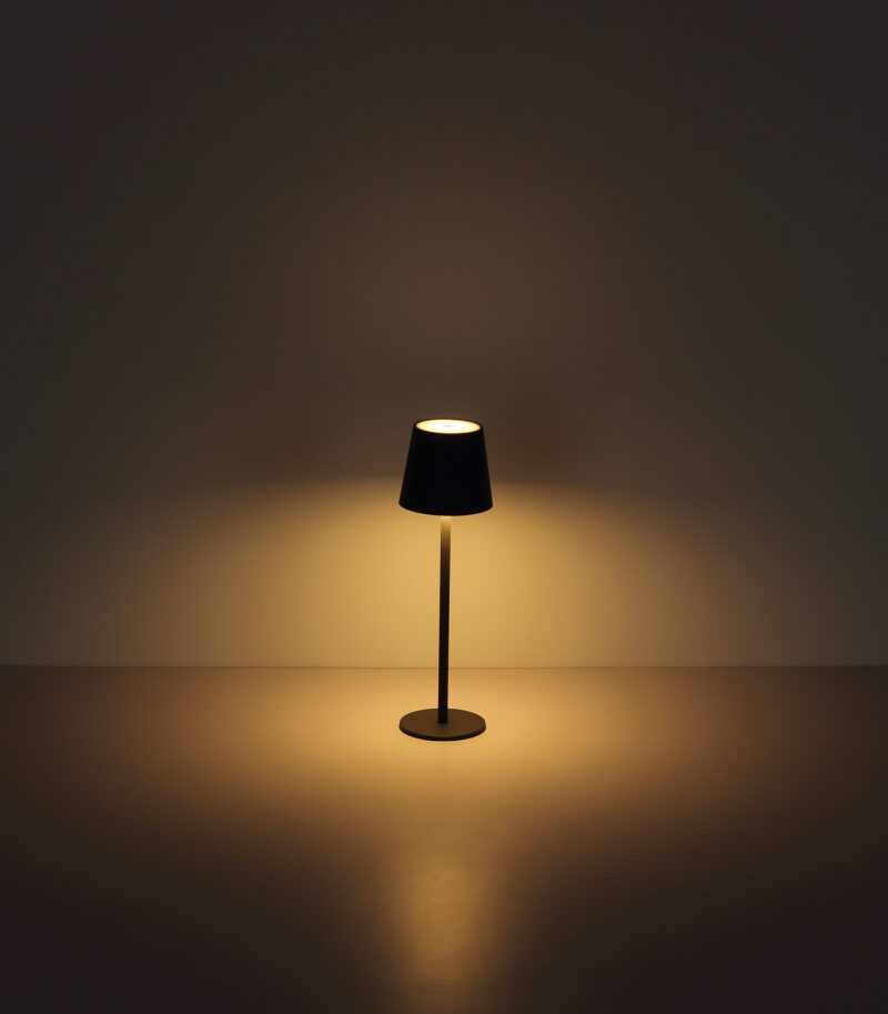 Die Tischleuchte - Vannie von Licht Factory ist eine moderne Stehleuchte mit dunklem Schirm, die ein warmes, sanftes Licht abgibt. Zentral vor einem schlichten Hintergrund platziert, schafft sie eine ruhige und minimalistische Atmosphäre.