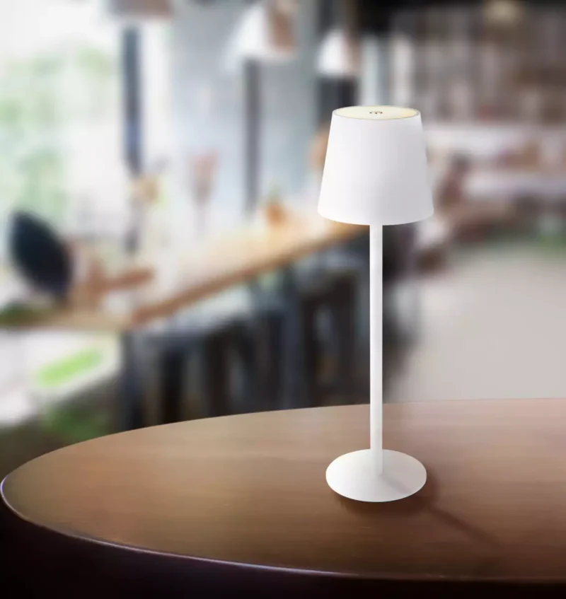 Die Tischleuchte - Vannie, eine elegante weiße Tischlampe von Licht Factory, verfügt über einen zylindrischen Sockel und einen runden Lampenschirm. Sie steht elegant auf einer Holzoberfläche vor einem verschwommenen Café-Hintergrund mit Tischen und Stühlen und schafft eine einladende Atmosphäre.
