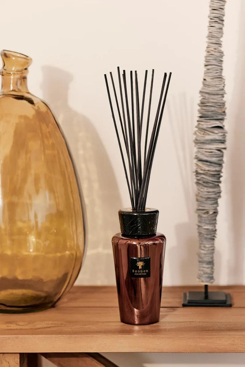 Der Raumduft - Exklusives Cyrium von Licht Factory, ein bronzefarbener Rattanstäbchen-Diffusor mit schwarzen Stäbchen, steht auf einer Holzoberfläche. Links davon steht eine große Vase aus bernsteinfarbenem Glas und rechts ein kunstvolles Stück gestapelter, unregelmäßig geformter Scheiben auf einem Ständer.