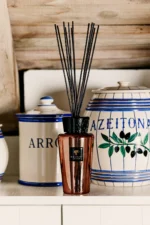 Der Raumduft - Exklusives Cyrium von Licht Factory, ein eleganter bernsteinfarbener Rattanstäbchen-Diffusor, ist stilvoll vor Keramikgefäßen mit den Aufschriften „Arroz“ und „Azeitona“ in blauen und grünen Designs platziert. Diese ruhige Präsentation auf einem weißen Regal vor einem hölzernen Hintergrund verbindet Tradition mit moderner Eleganz.