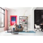 Eine Person mit rosa Haaren sitzt auf einem beigen Sitzsack in einem stilvollen Wohnzimmer von Licht Factory mit Marmorwänden. Das Gemälde „Wandbild - Pinky“, ein blauer Teppich, moderne Regale und runde Tische ergänzen den Raum. Große Fenster mit rosa Vorhängen lassen natürliches Licht herein.