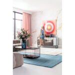 In einem modernen Wohnzimmer der Licht Factory sitzt eine Frau in einem blauen Kleid auf einem Sideboard. Der Raum ist mit abstrakter Kunst in „Circle Pink“ dekoriert, hat rosa Vorhänge, Blumen auf dem Couchtisch über einem blauen Teppich und große Fenster, die den Raum mit natürlichem Licht durchfluten.