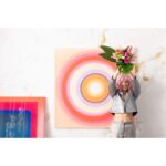 Eine Person mit rosa Haaren hält einen Blumenstrauß vor dem „Wandbild - Circle Pink“ hoch, einem farbenfrohen Gemälde mit konzentrischen Kreisen auf einer Wand mit Marmorstruktur. In der Nähe ergänzt ein weiteres Kunstwerk mit einem rosa-blauen Farbverlauf ihr Ensemble aus silberner Jacke und grauer Hose.