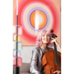 Eine Person mit rosa Haaren spielt Cello und lächelt, während ihre glänzende silberne Jacke das Licht einfängt. Dahinter harmoniert das lebendige „Wandbild - Circle Pink“ von Licht Factory perfekt mit einem Dekorationsobjekt aus gestapelten pastellfarbenen Würfeln.
