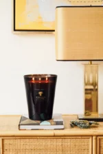 Eine große schwarze Duftkerze - Encre de Chine mit drei brennenden Flammen steht auf einem Holztisch neben einem Stapel Bücher. In der Nähe steht eine moderne Messinglampe von Licht Factory mit rechteckigem Schirm. An der Wand ist ein abstraktes Kunstwerk zu sehen.