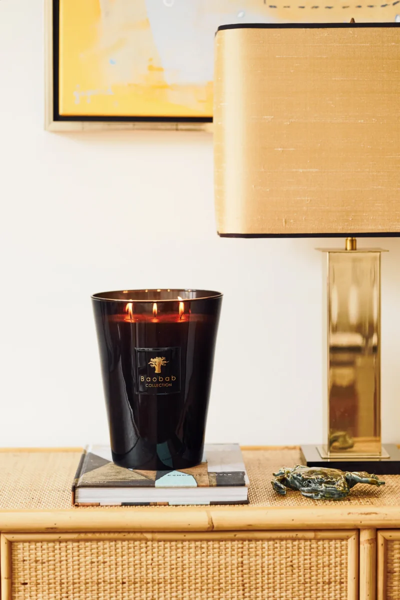 Eine große schwarze Duftkerze - Encre de Chine mit drei brennenden Flammen steht auf einem Holztisch neben einem Stapel Bücher. In der Nähe steht eine moderne Messinglampe von Licht Factory mit rechteckigem Schirm. An der Wand ist ein abstraktes Kunstwerk zu sehen.