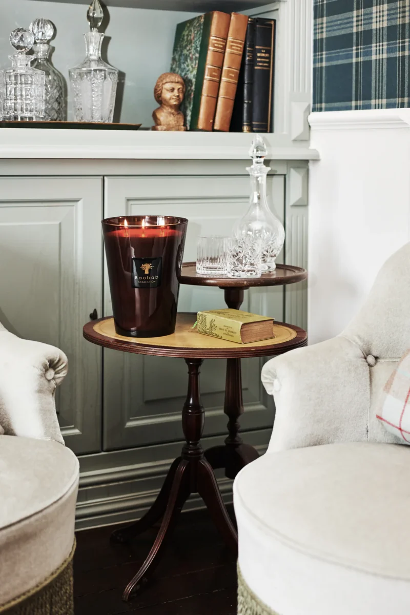 Ein gemütliches Interieur von Licht Factory präsentiert einen kleinen Beistelltisch aus Holz mit der Duftkerze - Encre de Chine, einer Glaskaraffe und Büchern. Ein weißer Schrank mit weiteren Büchern und Glaswaren ist der Mittelpunkt der Szene, während zwei elegante Polstersessel den Tisch flankieren.