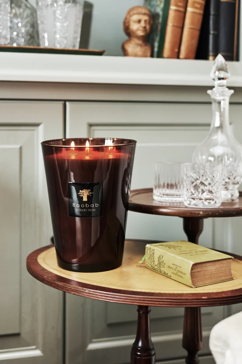 Die Duftkerze - Encre de Chine von Baobab Collection in einem durchscheinenden dunkelbraunen Glashalter steht elegant auf einem kleinen runden Tisch. In der Nähe schmücken ein Buch und eine Kristallkaraffe von Licht Factory einen größeren Tisch, alles eingerahmt von einem hellgrauen Schrankhintergrund.