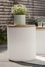 Auf einer Terrasse steht ein weißer zylindrischer Tisch von Licht Factory mit Holzplatte. Im Topf ruht eine grüne Pflanze im eleganten LED-Blumentopf von Lumi, geschmückt mit kleinen weißen Blüten. Daneben stehen zwei leere Gläser, eingerahmt von einer Außenszene mit einem Holzzaun und üppigem Grün.