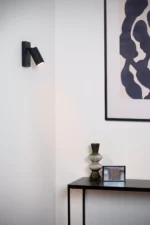 In einem modernen Interieur beleuchtet die Wandleuchte - Icon von Licht Factory eine weiße Wand, die mit abstrakter Kunst in einem schwarzen Rahmen geschmückt ist. Auf einem eleganten Konsolentisch stehen eine dekorative Glasvase und ein kleines gerahmtes Bild, was dem Raum Eleganz verleiht.