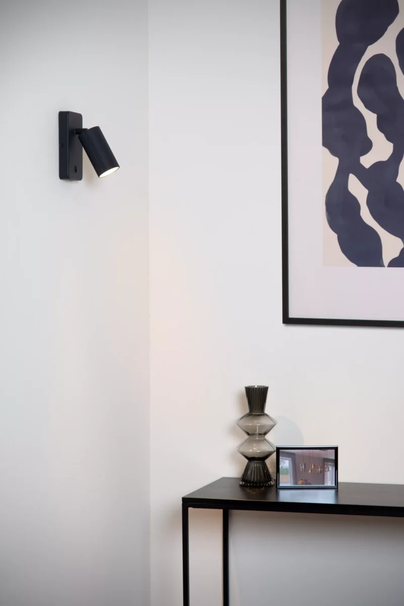 In einem modernen Interieur beleuchtet die Wandleuchte - Icon von Licht Factory eine weiße Wand, die mit abstrakter Kunst in einem schwarzen Rahmen geschmückt ist. Auf einem eleganten Konsolentisch stehen eine dekorative Glasvase und ein kleines gerahmtes Bild, was dem Raum Eleganz verleiht.