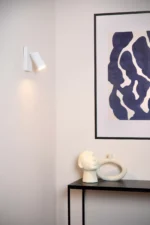 Das minimalistische Interieur von Licht Factory zeigt ein gerahmtes abstraktes Kunstwerk an einer weißen Wand. Die elegante Wandleuchte - Icon daneben wirft Licht auf einen schwarzen Tisch, der mit einem skulpturalen Kopf und einem ovalen Keramikstück geschmückt ist.