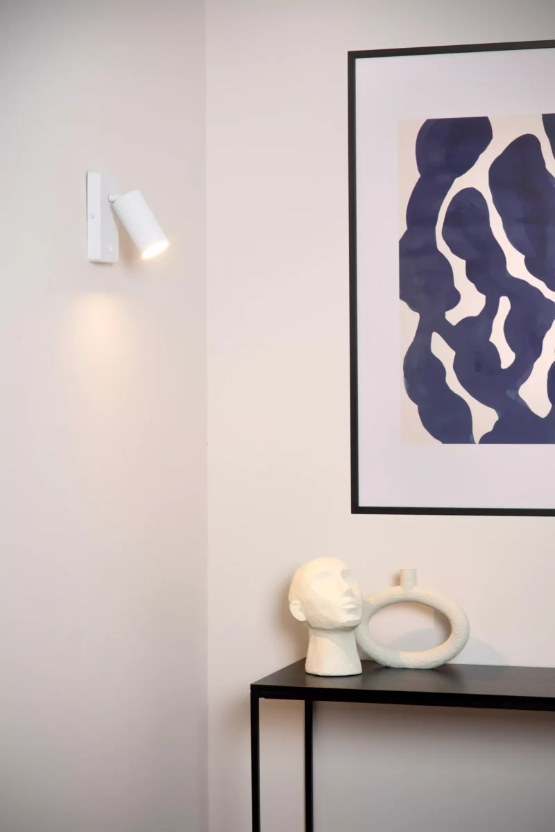 Das minimalistische Interieur von Licht Factory zeigt ein gerahmtes abstraktes Kunstwerk an einer weißen Wand. Die elegante Wandleuchte - Icon daneben wirft Licht auf einen schwarzen Tisch, der mit einem skulpturalen Kopf und einem ovalen Keramikstück geschmückt ist.