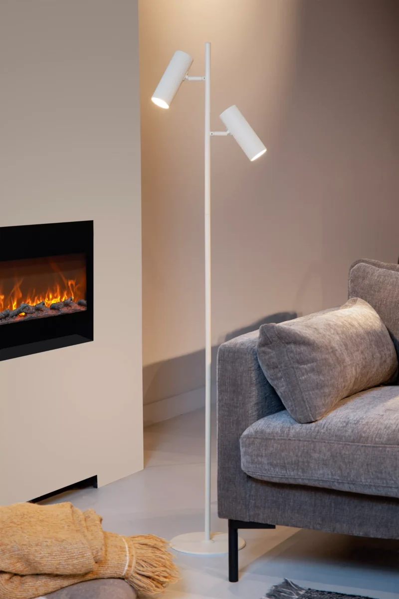 Ein gemütliches Wohnzimmer präsentiert die Stehleuchte - Icon von Licht Factory mit zwei zylindrischen Leuchten. Ein graues Sofa mit einer gelben Decke auf der Armlehne ist teilweise sichtbar und ein eingebauter Kamin leuchtet warm in der Wand.