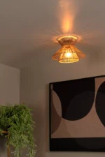 Die Deckenleuchte - Tassy von Licht Factory verbreitet mit ihrem geflochtenen Rattan-Design einen warmen Schein und erzeugt Deckenmuster. Sie beleuchtet ein großes gerahmtes abstraktes Kunstwerk in schwarzen und beigen Kreisformen an der Wand. Eine üppige grüne Pflanze lugt hindurch und wertet die Szene auf.