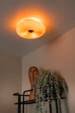 Die Deckenleuchte – Tino von Licht Factory wirft einen warmen Schein auf eine hölzerne Kopfskulptur und eine üppige grüne Pflanze auf einem modernen schwarzen Regal, während neutrale beige Wände die gemütliche Atmosphäre verstärken.