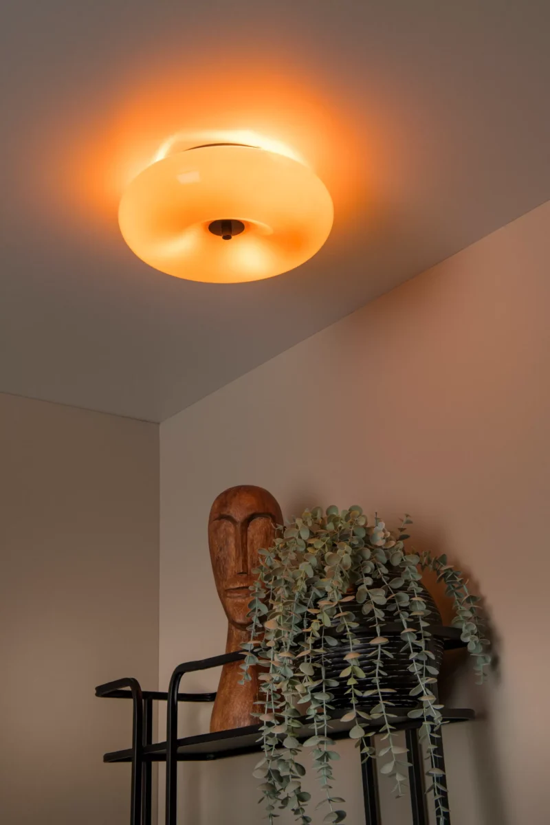 Die Deckenleuchte – Tino von Licht Factory wirft einen warmen Schein auf eine hölzerne Kopfskulptur und eine üppige grüne Pflanze auf einem modernen schwarzen Regal, während neutrale beige Wände die gemütliche Atmosphäre verstärken.