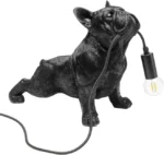 Die Tischleuchte - Toto hat die Form einer schwarzen, realistischen französischen Bulldogge mit detaillierten Merkmalen, die auf spielerische Weise eine Glühbirne im Maul hält und das Kabel von der Glühbirne aus verlängert.