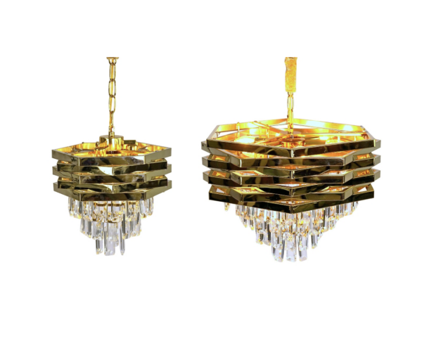 Zwei Kronleuchter aus Krystal Gold haben ein sechseckiges Design mit mehrschichtigen Metallringen und herabhängenden Kristallanhängern, die an Goldketten hängen und warmes Licht abgeben. Der Kronleuchter links ist etwas größer. Verfügbar ab Woche 6, 2025.