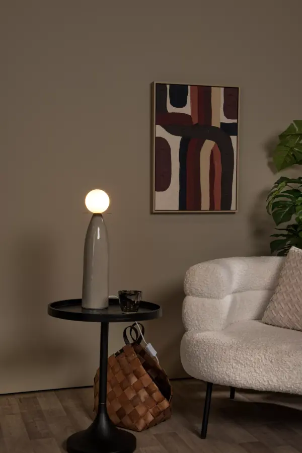 Ein gemütlicher Raum mit einem modernen weißen Sofa und einem runden schwarzen Beistelltisch, auf dem die Tischleuchte - Figaro steht, die neben einem Glas einen warmen Schein verbreitet. Darunter ergänzt ein geflochtener Korb das lebendige abstrakte Gemälde an der Wand. Eine grüne Pflanze haucht dem in neutralen Tönen gehaltenen Raum Leben ein.
