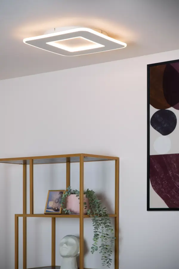 In einem modernen Raum ist die Deckenleuchte - Axell zu sehen, eine quadratische, minimalistische Deckenleuchte. Darunter befindet sich auf einem goldenen Metallregal eine kleine Topfpflanze, ein gerahmtes Foto und ein geformter weißer Kopf. An der Wand hängt ein geometrisches Kunstwerk mit Kreisen.