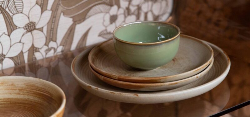 Eine grüne Keramiktasse steht auf einem Satz beiger Keramikteller auf einer reflektierenden Glasoberfläche. Im Hintergrund ist eine dekorative Wand oder Kulisse mit Blumenmuster zu sehen.
