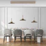 Ein modernes Esszimmer verfügt über einen langen Tisch für sechs Personen mit grau gepolsterten Stühlen, karierten Rückenlehnen und goldenen Akzenten. Darüber hängen drei minimalistische Pendelleuchten – PURE Moon Lights. Der Raum ist mit weiß getäfelten Wänden und einem Parkettboden geschmückt.