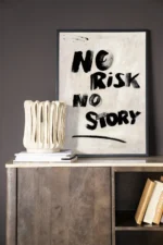An einer dunklen Wand hängt ein gerahmtes „Wandbild – No Risk, No Story“-Poster. Darunter steht eine strukturierte Keramikvase auf Büchern auf einem Holzschrank, auf dem untersten Regal lehnen weitere Bücher horizontal.