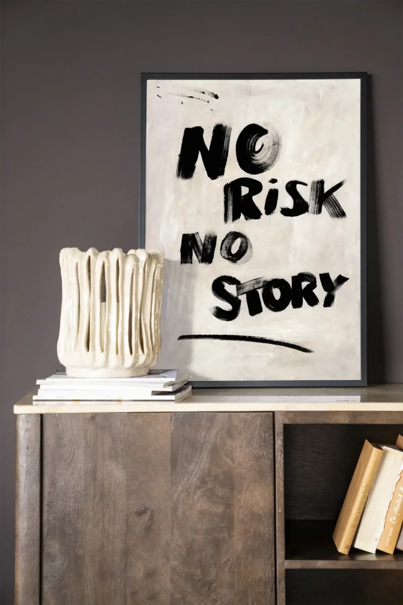 An einer dunklen Wand hängt ein gerahmtes „Wandbild – No Risk, No Story“-Poster. Darunter steht eine strukturierte Keramikvase auf Büchern auf einem Holzschrank, auf dem untersten Regal lehnen weitere Bücher horizontal.
