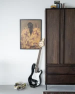 Eine schwarze E-Bassgitarre lehnt an einem Holzschrank, dazu gemusterte Turnschuhe. Darüber hängt ein gerahmtes Wandbild – Twins, das eine Person in einem gelben Hemd mit Blumenmuster zeigt. Auf dem Schrank liegen mehrere kleine Gegenstände.