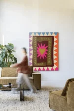 Eine Person geht an einem gemütlichen Wohnzimmer vorbei, in dem ein großes „Wandbild - Hug“ mit abstrakten Mustern und einer Sternform hängt, die das Motto „No Risk, No Story“ verkörpert. Im Zimmer stehen ein grüner Sessel, ein beiges Sofa, ein kleiner runder Tisch und eine Topfpflanze.