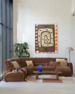 Ein modernes Wohnzimmer präsentiert ein großes braunes Sofa mit gemusterten Kissen, einen niedrigen hölzernen Couchtisch mit einer blauen Vase und eine große Pflanze neben raumhohen Fenstern. An der hellbeigen Wand hängt das farbenfrohe abstrakte Wandbild „Snake Eye“.