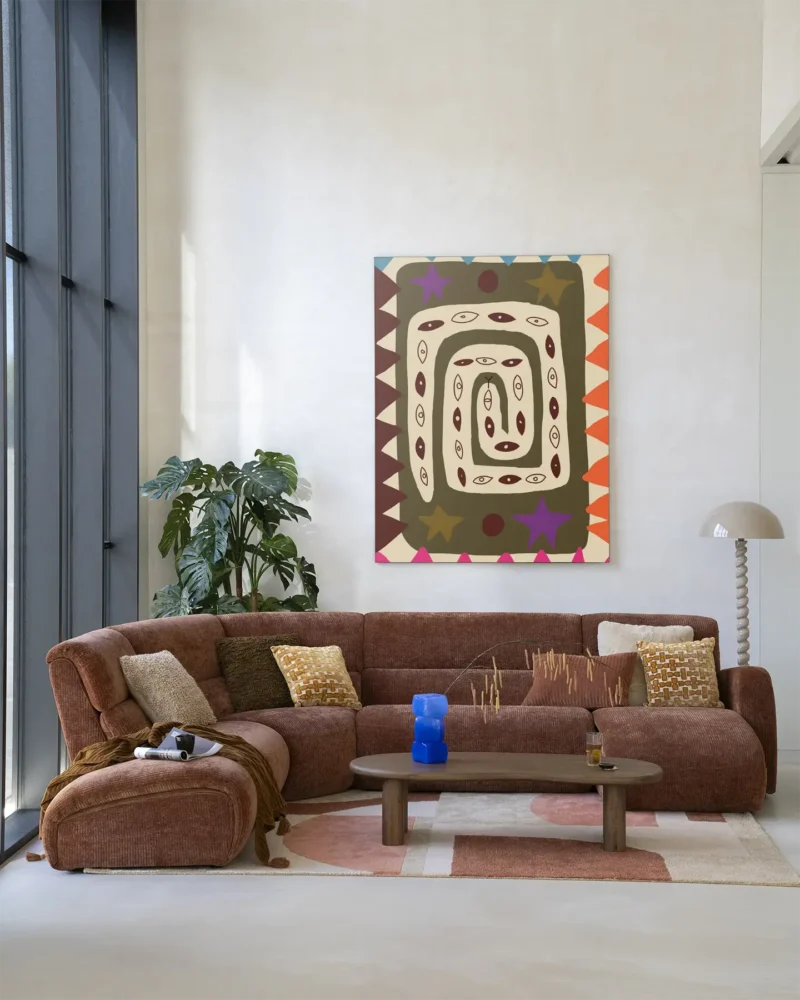 Ein modernes Wohnzimmer präsentiert ein großes braunes Sofa mit gemusterten Kissen, einen niedrigen hölzernen Couchtisch mit einer blauen Vase und eine große Pflanze neben raumhohen Fenstern. An der hellbeigen Wand hängt das farbenfrohe abstrakte Wandbild „Snake Eye“.