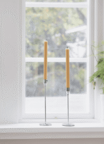 Zwei große, gelbe Kerzen in eleganten Koziol Kerzenhaltern - Siri zieren ein weißes Fensterbrett. Durch das Fenster lugt ein verschwommener Blick ins Grüne. Auf der rechten Seite des Fensterbretts ist teilweise eine Farnpflanze zu sehen.