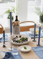 Ein Esszimmertisch besteht aus einer hellbraunen Tischdecke, blauen Servietten und hohen Kerzen in Siri-Kerzenhaltern. Eine zentrale Schüssel mit Muscheln ist von Gläsern, Tellern und einer in eine Serviette gewickelten Weinflasche umgeben.